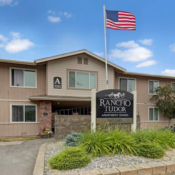 two-story apartment building with beige siding, marked "a 3531." a u.s. flag waves above the "rancho tudor apartment homes" sign amidst landscaping.