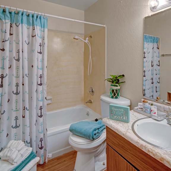 bathroom here at rancho tudor apartments with nautical-themed shower curtain featuring anchors, white bath, and sink with marble countertop. towel, plant, and decor add a beach vibe.