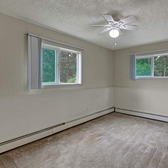 empty bedroom here at rancho tudor apartments with beige carpet and walls, featuring two large windows with vertical blinds. a ceiling fan with a light fixture hangs in the center.