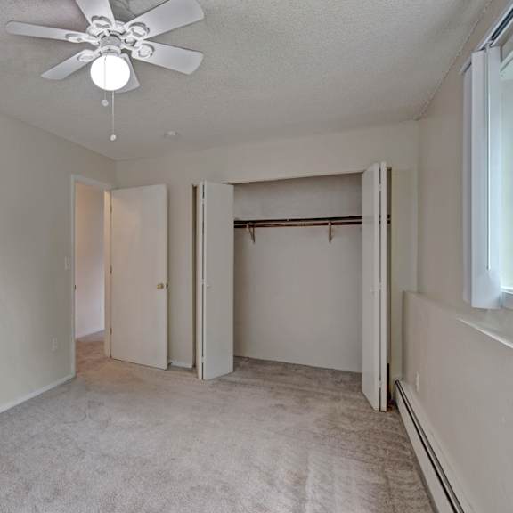 empty bedroom here at rancho tudor apartments with beige walls and carpet, featuring an open closet, a ceiling fan with light, and a window on the right letting in soft natural light.