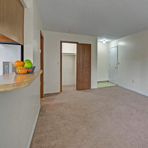 empty living room here at rancho tudor apartments with beige carpet and white walls, featuring a wooden door to a closet, a white entrance door, and a bar counter with a fruit bowl.