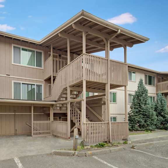 three-story beige rancho tudor apartment building with prominent wooden staircase and railings, set against a blue sky. nearby are parked bicycles and evergreen trees.