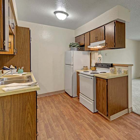cozy kitchen here at rancho tudor apartments with wooden cabinets, white appliances, and a laminate floor opens to a carpeted living area with a large window, offering natural light.