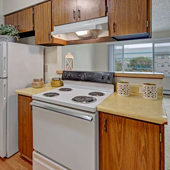 compact kitchen here at rancho tudor apartments with wood cabinets, white appliances including a stove and fridge. decor includes plants and lanterns, with ample natural light. cozy ambiance.