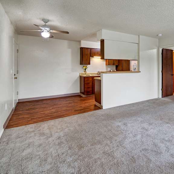 spacious empty living room here at rancho tudor apartments with beige carpet, white walls, and a small kitchen area with wooden cabinets. ceiling fan and open doorway add a welcoming feel.