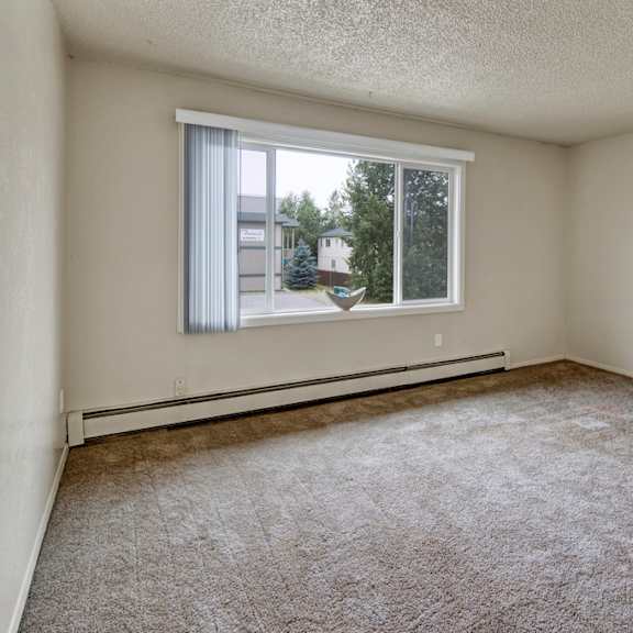 an empty beige room here at rancho tudor apartments with a large window showing trees and buildings outside. the carpet is brown, the walls are plain, and there's a minimal, neutral feel.