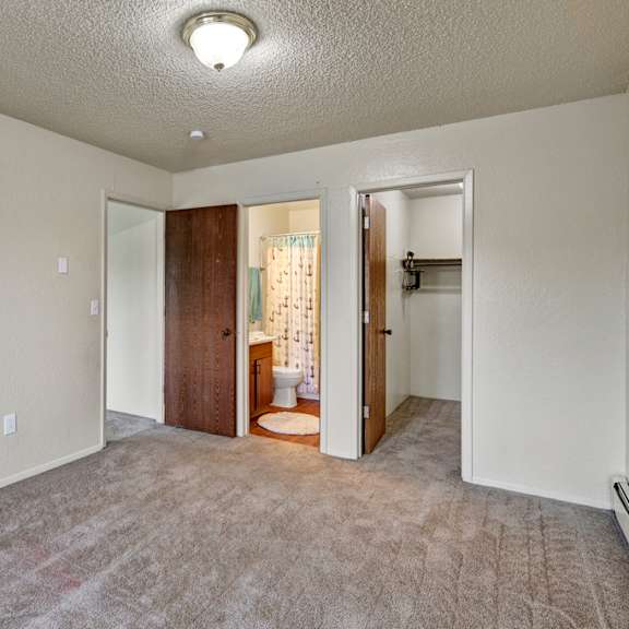 empty carpeted room here at rancho tudor apartments with beige walls, featuring two wooden doors. one door leads to a bathroom with a shower curtain; the other to a closet. a ceiling light softly illuminates the space.