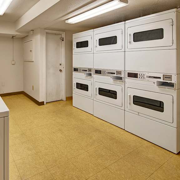 laundry room here at rancho tudor apartments with white machines and beige floor. six stacked dryers are on the right, and five washing machines are on the left. bright fluorescent lights illuminate the room.