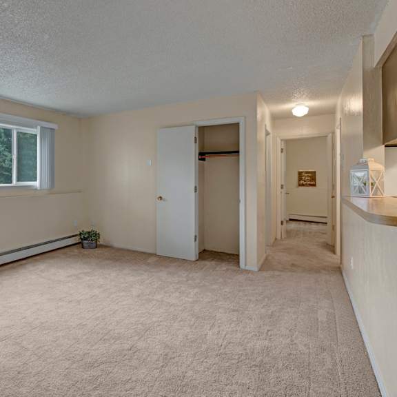 empty beige living room here at rancho tudor apartments with a large window showing greenery outside. the room features carpet, an open closet with a door, and a hallway with dim lighting. calm and neutral ambiance.