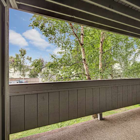 covered balcony here at russian jack apartments with wooden railing, beige carpet, and closed gray door, overlooking lush green trees. bright sky and distant buildings create a peaceful setting.