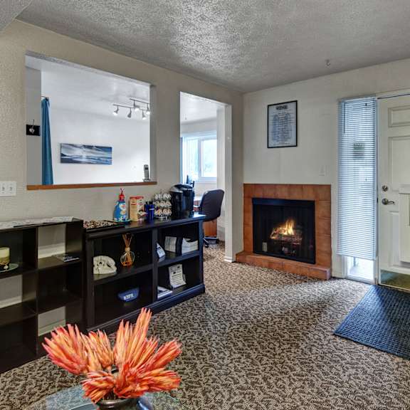 cozy office entrance here at russian jack apartments with a lit fireplace, shelf with snacks and supplies, and a large clock above a white door flanked by blinds. warm ambiance.