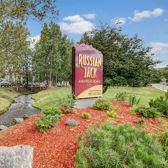 sign for russian jack apartment homes by a small creek with red mulch landscaping, surrounded by lush trees and a clear blue sky, creating a peaceful setting.