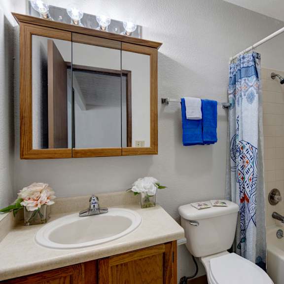 small, tidy bathroom here at russian jack apartments with a white sink, wooden cabinet, and mirror. a flower arrangement and blue towels add color. shower has a patterned curtain.