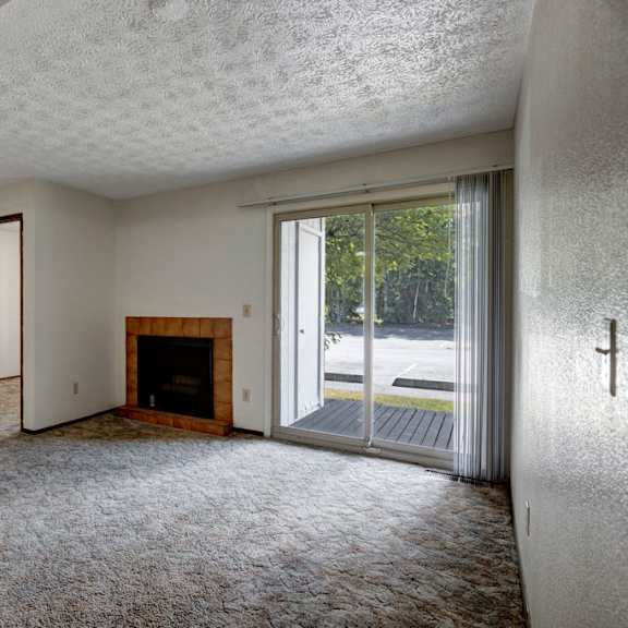 empty living room here at russian jack apartments with textured ceiling, beige carpet, and neutral walls. features a brick fireplace, sliding glass door with vertical blinds, and natural light. atmosphere feels calm and open.