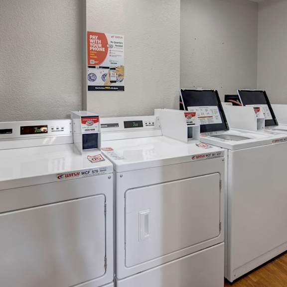 a row of white commercial washers and dryers is in a clean, gray laundry room here at russian jack apartments. a poster on the wall advertises mobile payment options.