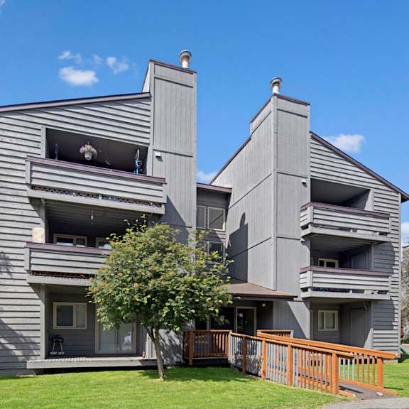 gray three-story russian jack apartment building with balconies, set against a backdrop of blue sky and trees. a wooden ramp leads to the entrance. calm atmosphere.