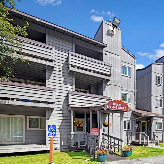 gray, multi-story russian jack apartment building with balconies under a blue sky. a leasing office sign is visible. accessible parking signs are in front.