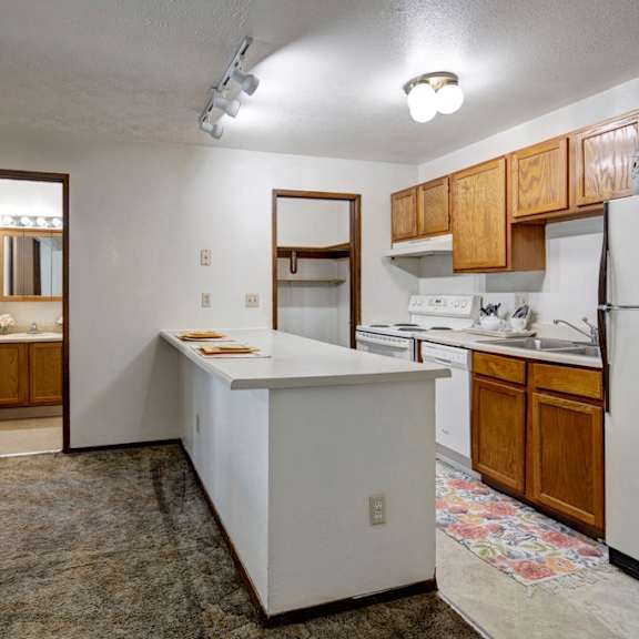 compact kitchen here at russian jack apartments with wooden cabinets, white appliances, and a floral rug. a long countertop extends into a hallway showing a lit bathroom mirror. cozy feel.