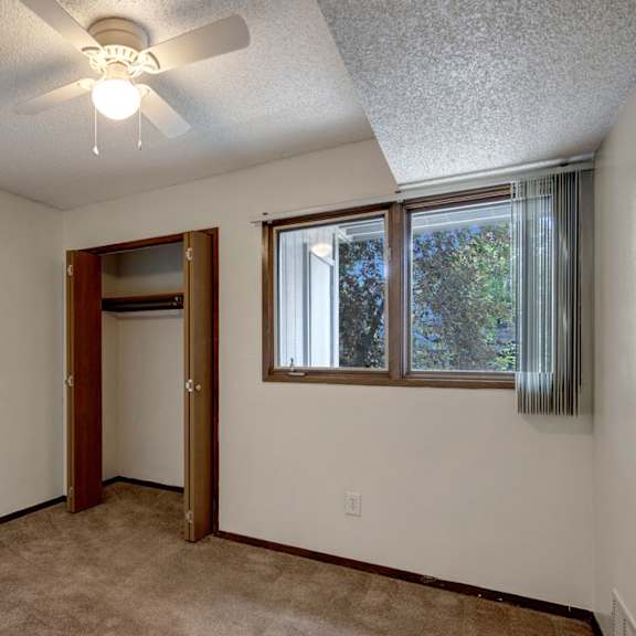 small, empty bedroom here at russian jack apartments with beige carpet, white walls, and a ceiling fan. open closet with wooden doors and a window on the right with vertical blinds.