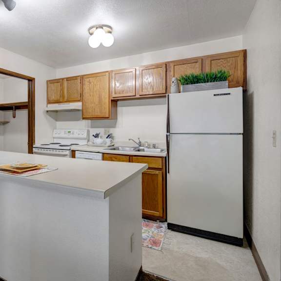 small, cozy kitchen here at russian jack apartments with overhead lighting, featuring wooden cabinets, a white fridge, stove, and sink. simple counter with two place settings.