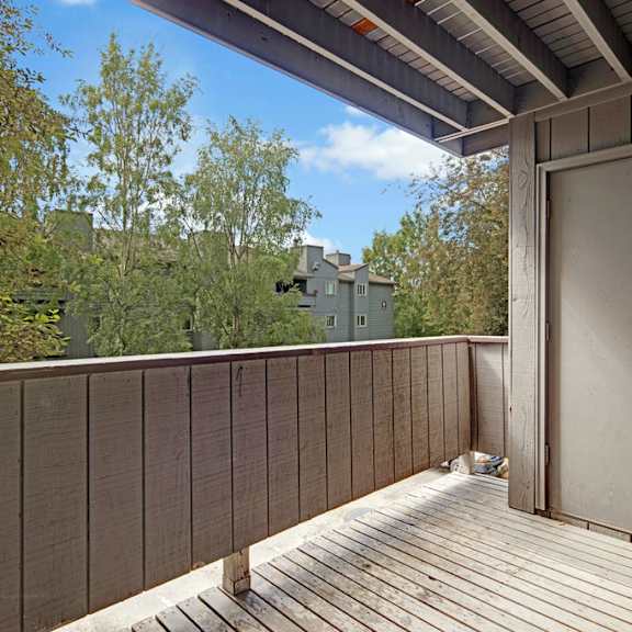 a wooden balcony here at russian jack apartments with railing offers a view of trees and distant buildings. a closed door is on the right, under a partially covered roof, in daylight.