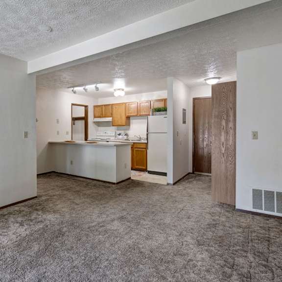 empty living room here at russian jack apartments with beige carpet, white walls, and an adjacent kitchen featuring wooden cabinets, white appliances, and a small island. cozy and neutral.