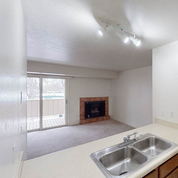small, empty living room here at russian jack apartments with beige walls and carpet. features a fireplace, sliding glass door to a balcony, and a kitchen view with a double sink. neutral lighting.
