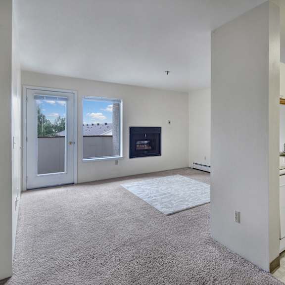 cozy apartment interior here at silver ridge apartments with carpeted living room, fireplace, and bright window. adjacent kitchen has white appliances and cabinets, and a warm tone.