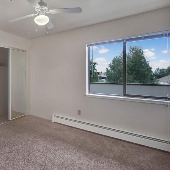 empty bedroom here at silver ridge apartments with beige carpet and white walls. large mirrored closet doors reflect a ceiling fan. bright window view reveals green trees and blue sky.
