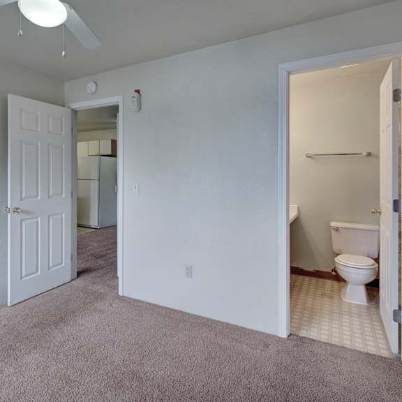 empty bedroom here at silver ridge apartments with gray walls and carpet, open door to a bathroom with a toilet and towel rack. ceiling fan overhead. doorway reveals a kitchen area with a fridge.
