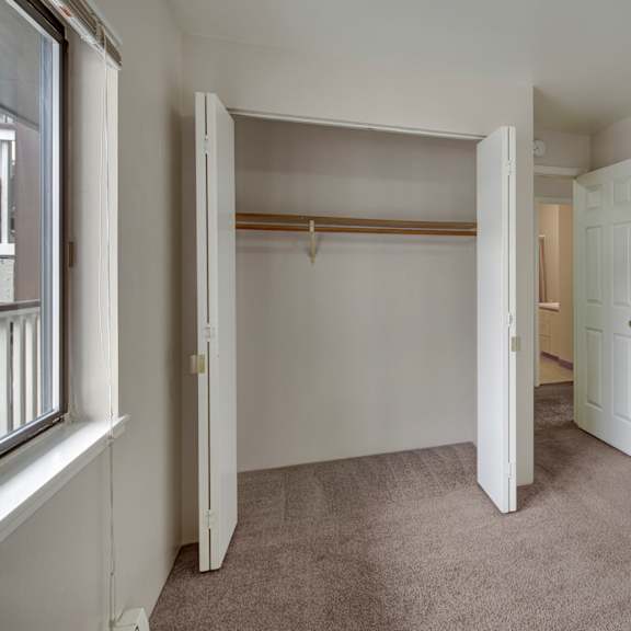 empty bedroom here at silver ridge apartments with beige carpet, open closet with wooden rod, white walls, and doors leading to another area. a window on the left brings in light.