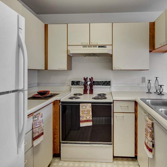 compact kitchen here at silverr ridge apartments with white cabinets and countertops, electric stove, refrigerator, and double sink. decorative towels and figurines add warmth.