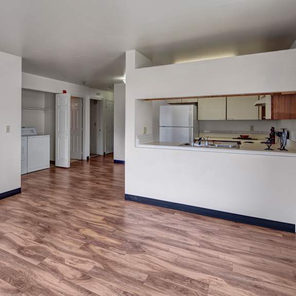spacious, empty living room here at silver ridge apartments with light wood flooring and white walls. open kitchen with a visible fridge and countertop, providing a clean, minimalist feel.
