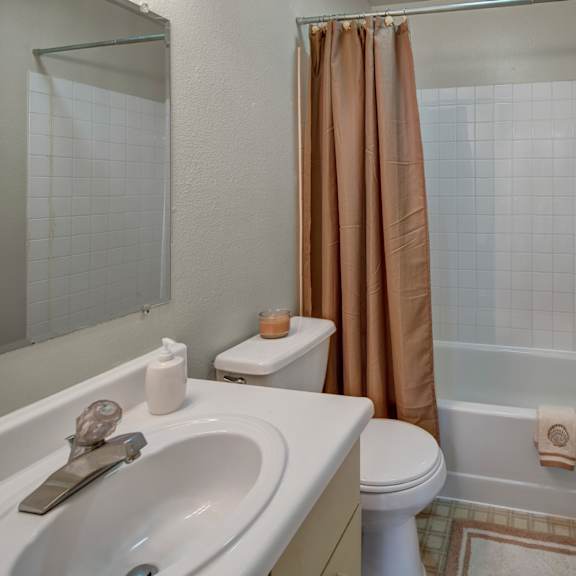 a small bathroom here at silver ridge apartments with white sink and toilet, beige shower curtain, and matching towels with shell motifs. a candle adds a warm, inviting touch.