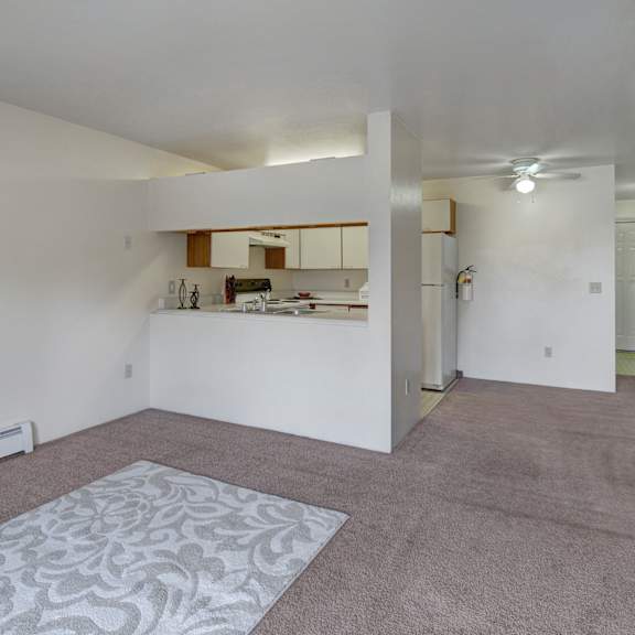 spacious living room here at silver ridge apartments with beige carpet and patterned rug. open view of a white kitchen with appliances. ceiling fan and soft lighting create a cozy atmosphere.