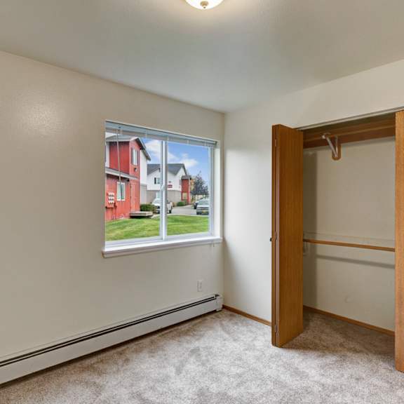 small, empty room here at strawberry lane apartments with beige walls and carpet, open wooden closet doors, and a window showing green grass and red buildings outside, creating a calm tone.