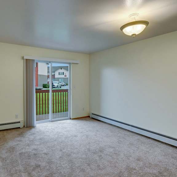 empty beige room here at stawberry lane apartments with carpet, bright ceiling light, and sliding glass doors leading to a small balcony overlooking grass and residential houses. calm ambiance.