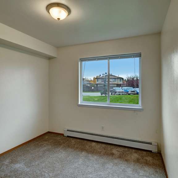 small empty room here at strawberry lane apartments with beige carpet and cream walls, featuring a ceiling light and a window overlooking parked cars and greenery, conveying a simple, quiet atmosphere.