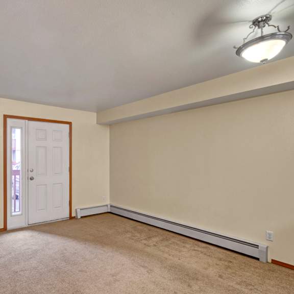 empty living room here at strawberry lane apartments with beige walls and carpet, featuring a white door with a side window. a round ceiling light casts a soft glow, creating a serene atmosphere.