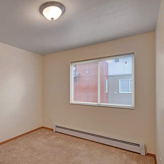 small, empty beige room here at strawberry lane apartments with carpeted floor, single window with blinds, and a ceiling light. the room has a simple, minimalist appearance.