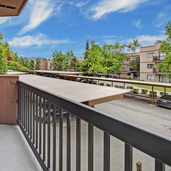 a small balcony here at susitna ridge apartments with a metal railing overlooks a parking lot and covered parking. surrounding are beige apartment buildings and green trees under a blue sky.