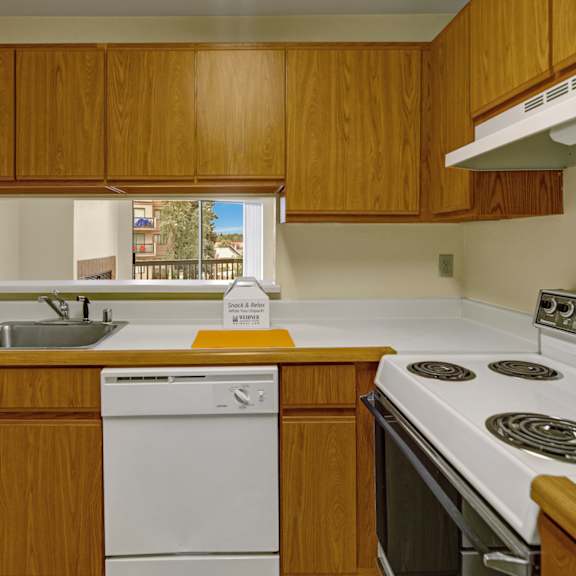 compact kitchen here at susitna ridge apartments with wooden cabinets, white appliances, and a stainless steel sink. a window view of trees and buildings adds warmth to the space.