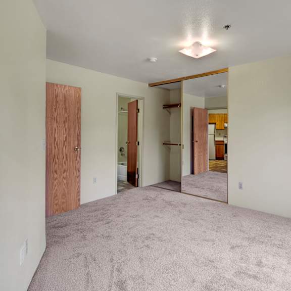 empty room here at susitna ridge apartments with beige carpet, mirrored closet doors reflecting a kitchen, wooden doors leading to a bathroom; minimalistic and bright atmosphere.