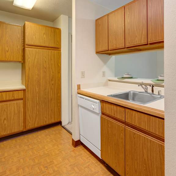 compact kitchen here at susitna ridge apartments with brown wooden cabinets, white countertops, and a sink. a glimpse of a carpeted living area with sliding glass doors to a balcony. cozy atmosphere.