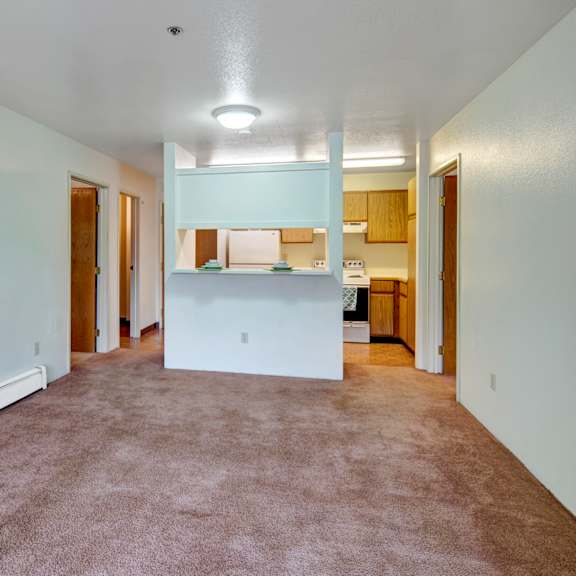 spacious, empty apartment living room here at susitna ridge apartments with beige carpet and white walls, a hallway with wooden doors, and a small kitchen with light wood cabinets.