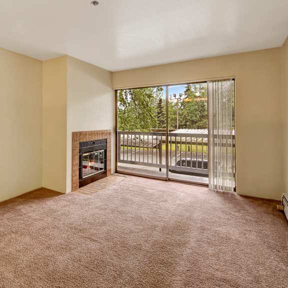 empty living room here at susitna ridge apartments with beige carpet and walls, featuring a tiled fireplace. a large sliding glass door leads to a balcony with views of greenery. bright and serene atmosphere.