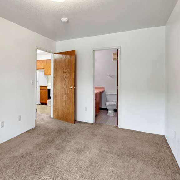 empty bedroom here at susitna ridge apartments with beige carpet and white walls, featuring open wooden doors leading to a kitchen with wooden cabinets and a bathroom. bright, neutral tone.