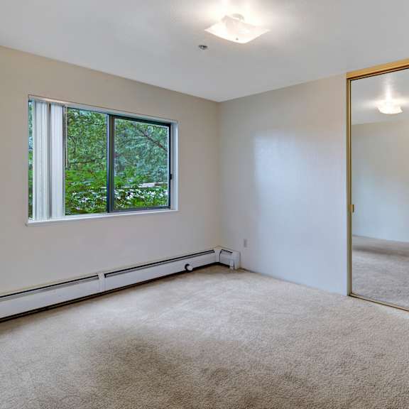 empty, bright bedroom here at susitna ridge apartments with beige carpet, large window showing greenery, and mirrored sliding closet doors reflecting ceiling light, creating a serene atmosphere.