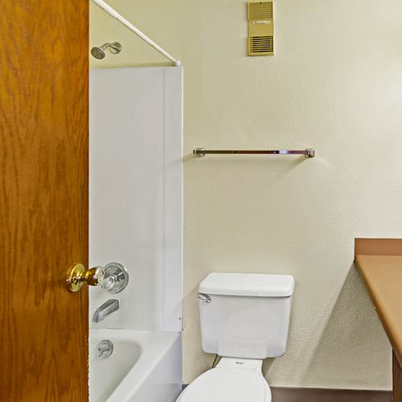 compact bathroom here at susitna ridge apartments with a white tub, shower, and toilet. brown countertop with a sink and large mirror. beige walls create a neutral, minimal tone.