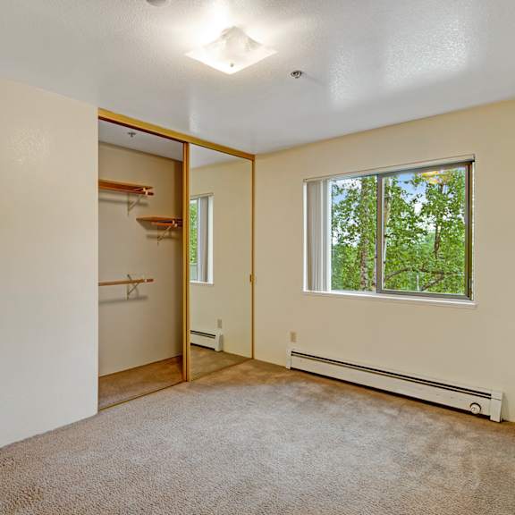 empty beige-walled bedroom here at susitna ridge apartment with carpet, a mirrored closet door, and a window revealing green trees outside. bright and simple, evokes a calm atmosphere.